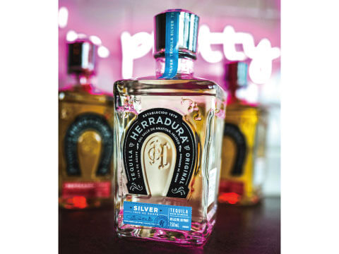 A bottle of Herradura Silver tequila featuring its horseshoe logo, displayed against a pink-lit bar backdrop