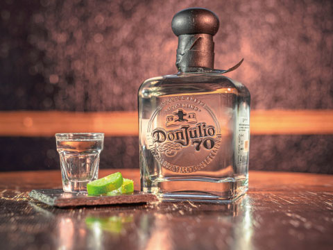 A bottle of Don Julio 70 blanco tequila beside a filled shot glass and fresh lime wedges, set on a dark wooden bar table
