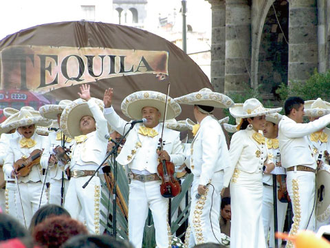 A mariachi band in traditional white and gold charro suits performing with violins outdoors, with a large "Tequila" banner in the background