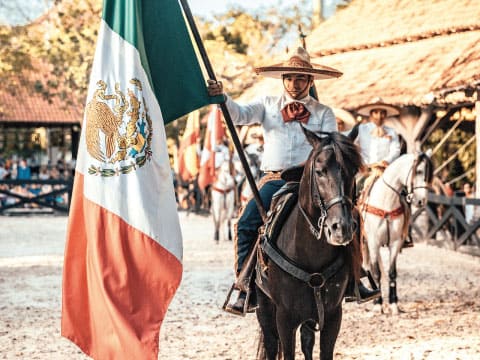 A Mexican charro horseman wearing a wide-brimmed sombrero rides a dark horse while holding the Mexican flag, with fellow riders and a traditional hacienda-style building in the background