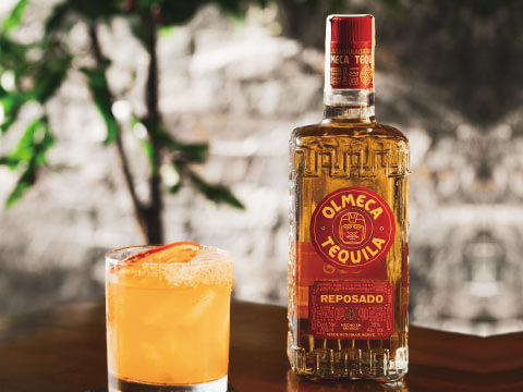 A bottle of Olmeca Reposado tequila beside a salt-rimmed orange cocktail garnished with citrus, set on a wooden table with greenery in the background