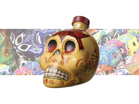 A hand-painted skull-shaped bottle of KAH Reposado tequila featuring a red bandana design, set against a colorful background of Day of the Dead sugar skull artwork
