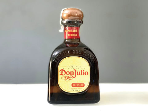 A bottle of Don Julio Reposado tequila with its short, squat silhouette and cork-topped cap, photographed against a plain gray background