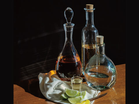 Three tequila varieties — dark, gold, and clear — in glass decanters beside a shot glass and lime wedges