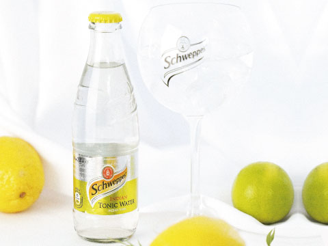 A bottle of Schweppes Indian Tonic Water beside an empty branded glass, surrounded by fresh lemons and limes on a white background