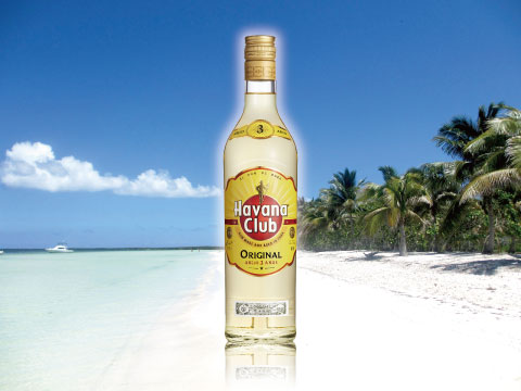 A bottle of Havana Club Original 3 Años rum standing on a white sand beach with turquoise water and palm trees
