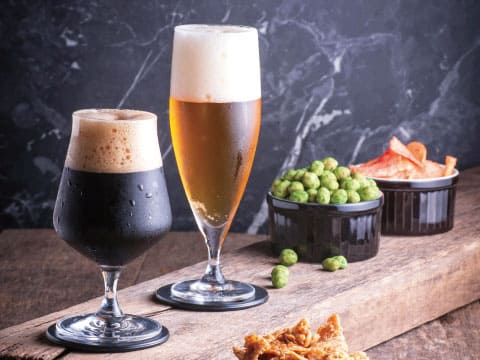 A dark stout and a golden lager in glasses on coasters, beside bowls of nuts and chips on a wooden table