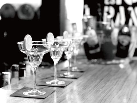 A black-and-white photo of several salt-rimmed margarita glasses lined up along a wooden bar counter, garnished with lime wheels
