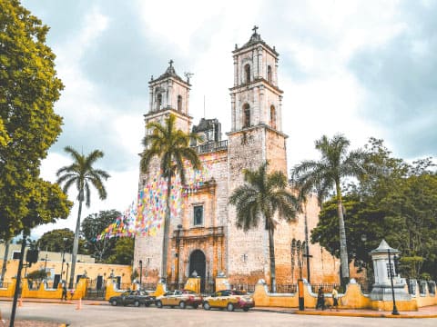 The twin-towered San Servacio Cathedral in Valladolid, Yucatán, with palm trees and colorful papel picado banners in front