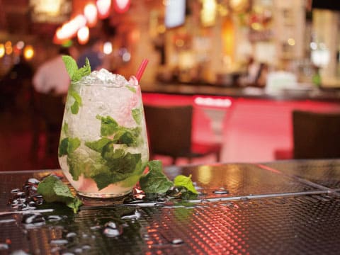 A mojito cocktail garnished with mint leaves and a straw, sitting on a wet bar table with blurred bar lights in the background