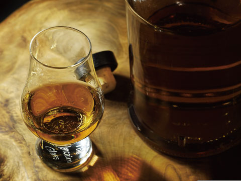 A glass of amber gold rum on a wooden barrel top, next to a large glass bottle