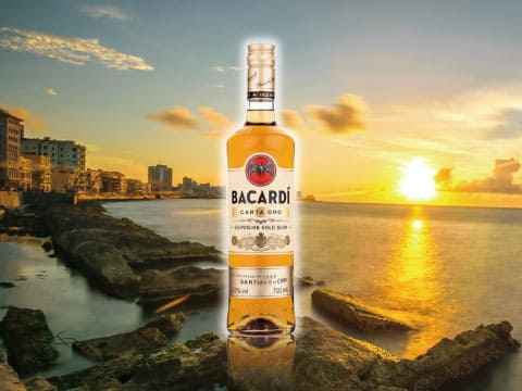A bottle of Bacardí Carta Oro gold rum standing on rocks along the Havana coastline at sunset