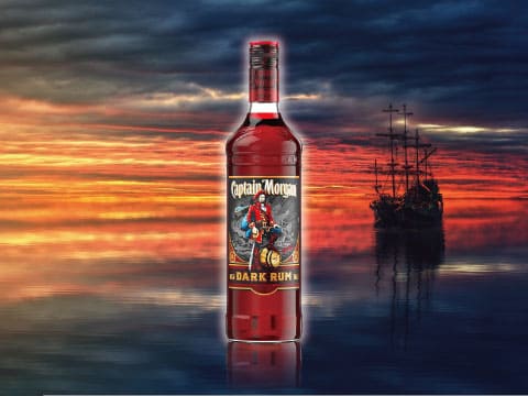 A bottle of Captain Morgan Dark Rum standing over calm water at sunset, with a pirate ship silhouette in the distance