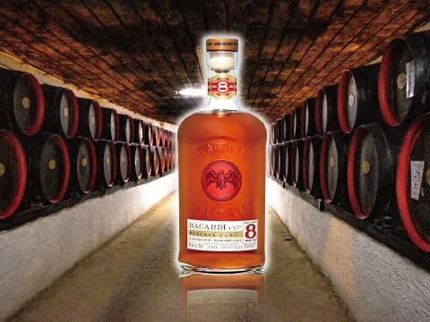 A bottle of Bacardí Ocho rum standing in a cellar corridor lined with rows of oak aging barrels