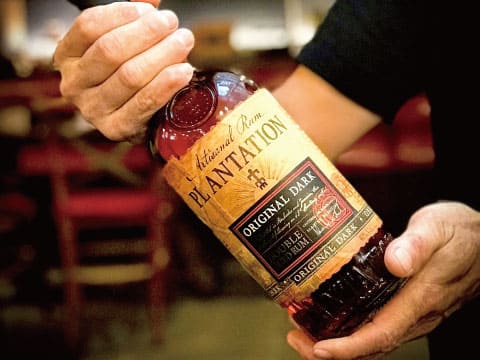 Two hands holding a bottle of Plantation Original Dark rum in a dimly lit bar setting