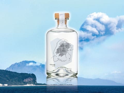A bottle of Acoua Rum White standing against a backdrop of coastal mountains and blue water