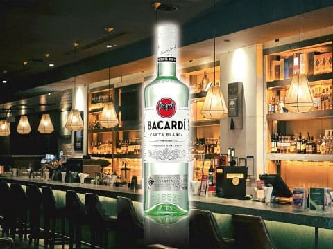 A bottle of Bacardí Carta Blanca white rum displayed against a blurred bar counter with warm pendant lighting