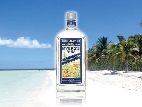A bottle of Myers's Rum Platinum White standing on a white sand beach with turquoise water and palm trees