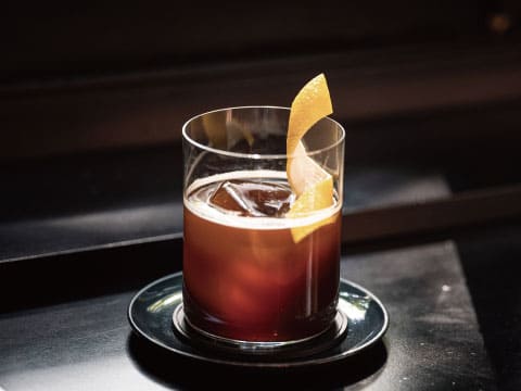 A dark brown cocktail served over ice in a rocks glass, garnished with a curled orange peel, placed on a black coaster