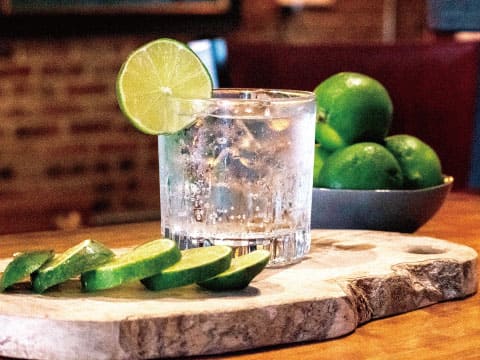 A clear beverage served over ice in a rocks glass, garnished with a lime wheel, placed on a wooden board with sliced limes and a bowl of whole limes nearby
