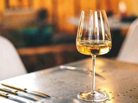 A glass of golden aperitif wine sits on a restaurant table beside cutlery, with warm ambient lighting in the background.