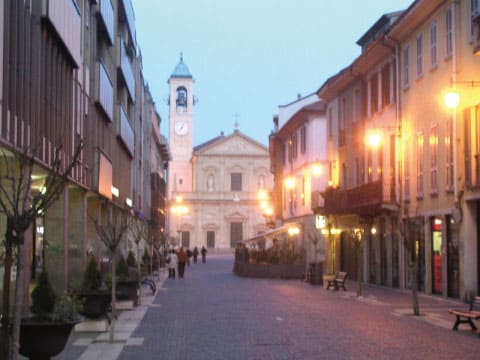 A cobblestone street in Saronno, Italy at dusk, lined with shops and a church bell tower in the background