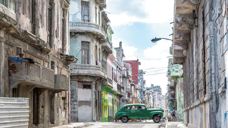 A narrow street in Old Havana lined with weathered colonial buildings and a vintage green car parked in the middle