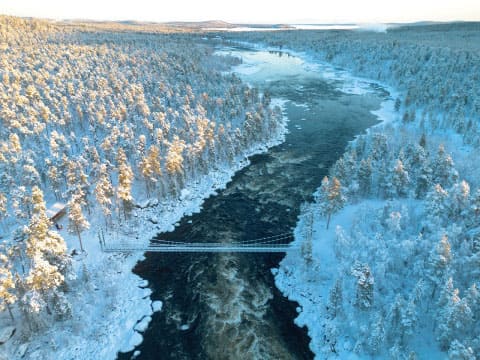 Aerial view of a dark river winding through a snow-covered forest valley with a suspension bridge in Finland