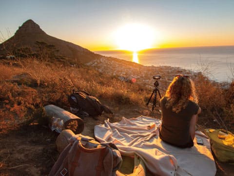 Woman with curly hair sitting on a picnic blanket photographing a sunset over a coastal city and ocean from a hilltop, with camera equipment and backpacks nearby