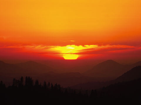 A fiery orange and red sunset with the sun setting behind silhouetted mountain ridges and trees