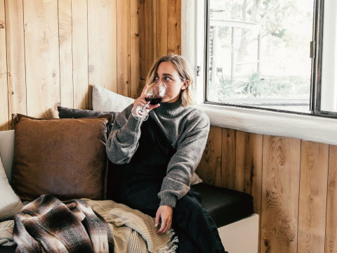 A woman in a gray sweater sipping red wine on a couch beside a window in a cozy wood-paneled room