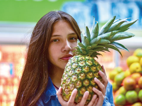A young woman holding a whole fresh pineapple close to her face, with colorful fruit blurred in the background