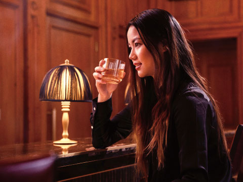A woman sitting at a bar counter, smiling while holding a glass of whisky, with a lamp glowing beside her