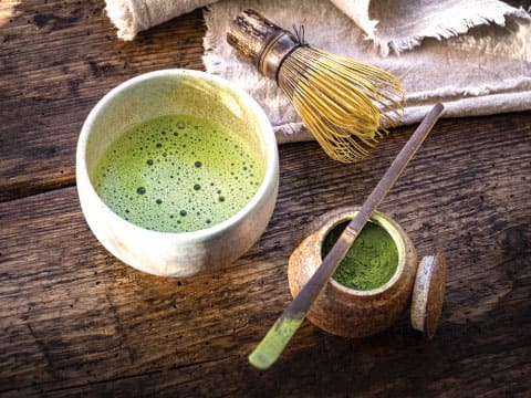 A bowl of frothy matcha tea sits beside a bamboo whisk, powder container, and scoop on a wooden table.