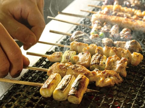Hands turning skewers of grilled chicken and green onion yakitori over a charcoal grill