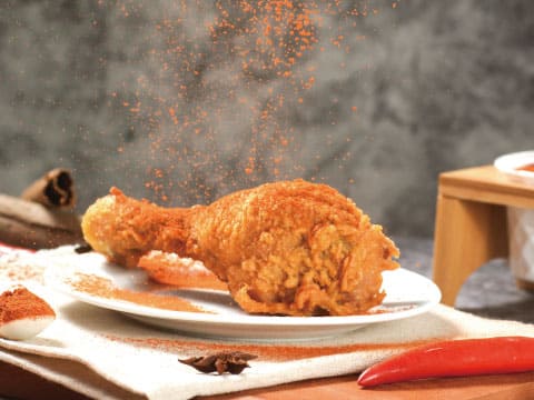 A crispy fried chicken drumstick dusted with red spice powder on a white plate, garnished with a red chili pepper, star anise, and cinnamon sticks on a wooden table