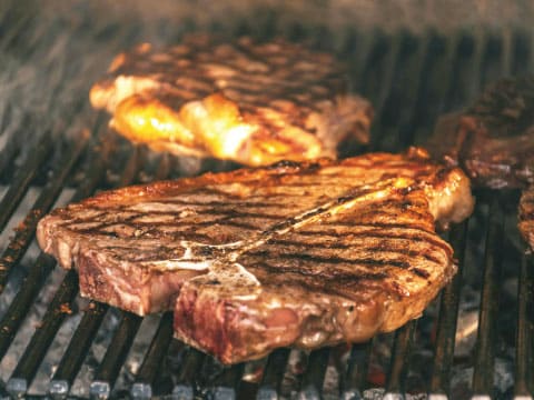 T-bone steak with grill marks cooking over an open flame grill, with grilled meat in the background