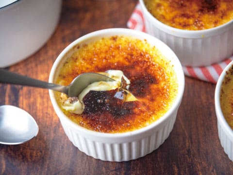 A close-up of crème brûlée in a white ramekin with a cracked caramelized sugar crust, a spoon dipped into the custard, with more ramekins in the background