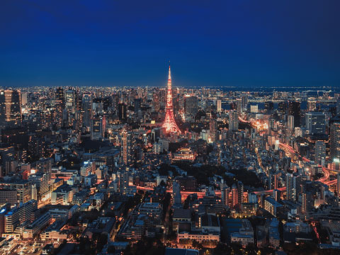 An aerial night view of Tokyo's cityscape with the illuminated red Tokyo Tower rising above the skyline, surrounded by city lights