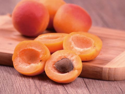 Whole and halved fresh apricots, one still with its pit, sit on a wooden cutting board.