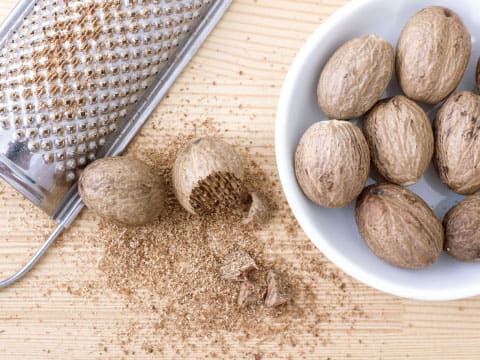 Whole nutmeg seeds sit beside a metal grater with freshly grated nutmeg powder scattered on a wooden surface.