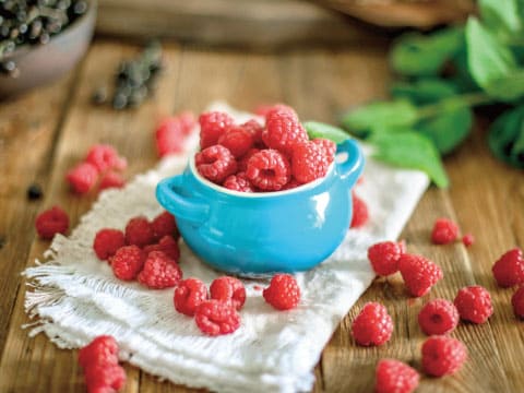 Fresh raspberries fill a small blue bowl, with more scattered around it on a wooden table.