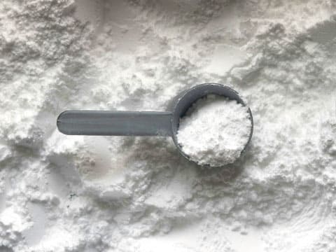 A gray measuring scoop holds fine white powder against a background of more scattered powder.