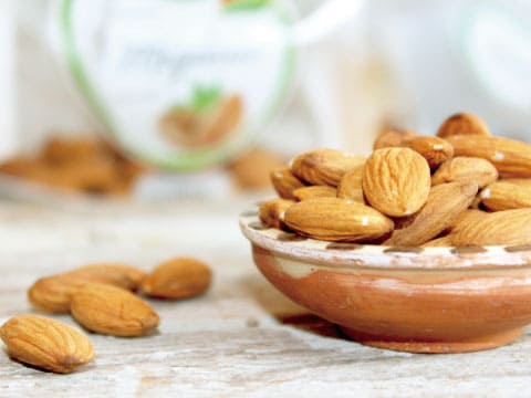 A small terracotta bowl filled with raw almonds, with more scattered on a wooden surface nearby
