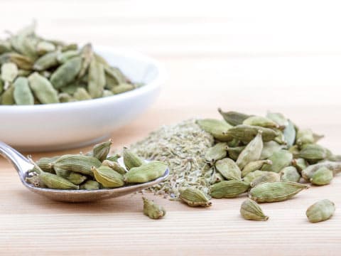 Whole green cardamom pods are piled on a spoon and scattered on a wooden table beside a bowl of more pods.