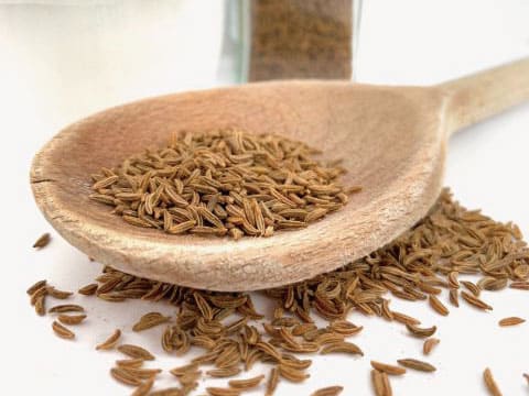 Whole caraway seeds are piled on a wooden spoon with more scattered on a white surface.