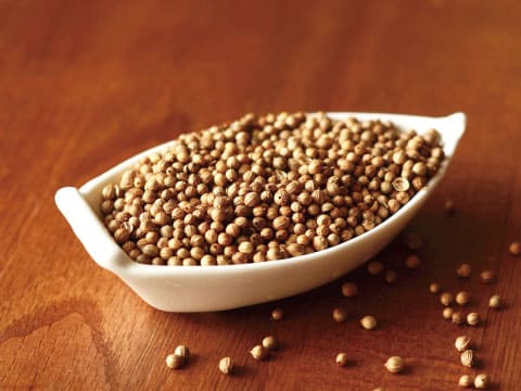 Dried coriander seeds fill a boat-shaped white bowl, with a few scattered on the wooden table beside it.