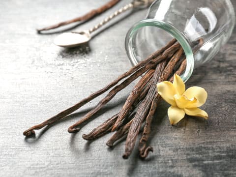 Dried vanilla beans spill out of a glass jar beside a vanilla flower and a spoonful of sugar on a wooden table.