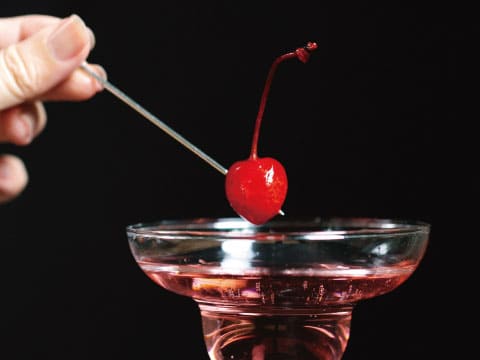 A hand places a bright red maraschino cherry on a cocktail pick above a pink drink in a coupe glass.