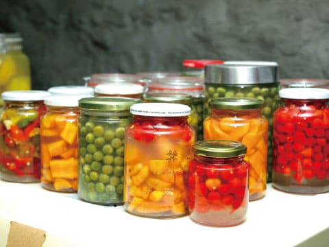 Rows of glass jars filled with preserved fruits and vegetables, including peas, peppers, tomatoes, and citrus, arranged on a table
